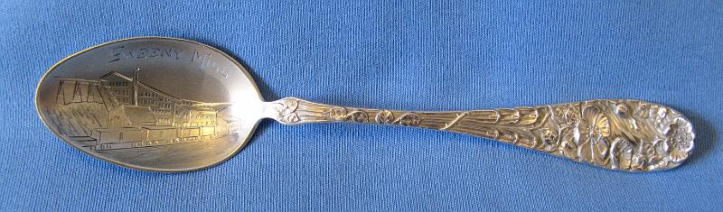 Souvenir Mining Spoon Sweeny Mill Wardner ID.JPG - SOUVENIR MINING SPOON SWEENY MILL WARDNER ID - Sterling silver spoon withengraved bowl showing mine buildings and marked SWEENY MILL with figural handle of flowers, 5 3/4 in. long, reverse marked Sterling with maker’s hallmark  (The Coeur d'Alene mining region in northern Idaho is one of the nation's most significant mineral producers, having yielded almost twenty percent of the silver and lead, and six percent of the zinc produced in the United States.  Due to the relatively low-grade of many of the lead-zinc-silver ores mined, early methods of concentration were introduced at the beginning of commercial mining operations in 1886. These milling operations produced large tonnages of mill tailings, the material remaining after crushing, grinding of the ore and separation of a significant portion of the economic minerals as a waste product.  The Sweeny or Last Chance mill located about two miles west of Kellogg, ID in the Coeur d’Alene District was a major contributor to the production of lead and silver as well as tailings.  In 1886 Charles Sweeny and F. R. Moore of Spokane opened the Last Chance mine, located near Wardner.  In May 1898, the Empire State-Idaho Mining and Development Company was organized to control the Last Chance and to acquire new territory to the west. In September 1903, the Federal Mining and Smelting Company was organized and purchased the Empire State holdings.  The Last Chance mine was a steady producer of lead-silver ore since 1890.  With the completion of the Oregon Railway and Navigation Company's Wallace branch in 1890, Charles Sweeny built a mill with a capacity of 500 tons daily to process ore from the Last Chance mine.  At the time, nearly 300 men were employed at the mine and mill.  The Last Chance mine was worked through a tunnel nearly two miles long that perforated the ridges west of Wardner at an elevation of 3,050 feet. One portal was called the Sweeny tunnel and the other the Arizona. Concentrating ore was taken from the Arizona tunnel over the Oregon Railway and Navigation track to the Sweeny mill at the mouth of Government gulch. Lingering litigation between the Last Chance Mining Company and its successors on the one hand and the Bunker Hill and Sullivan Company on the other over ownership of the deposits was ultimately decided in favor of the Bunker Hill and Sullivan Company.  The Last Chance mine and the Sweeny mill became the property of Bunker Hill & Sullivan Mining Co. in 1918.  Mitigation of heavy metals-bearing, tailings-contaminated river sediments continues as an issue in the region today since these sediments first reached farms and communities down river from the mines around 1900.)